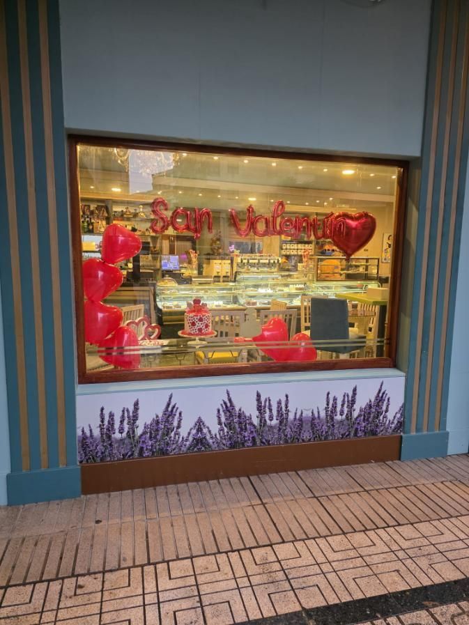 Escaparate de panadería con decoraciones de San Valentín, globos rojos y pancarta, productos horneados en el interior.