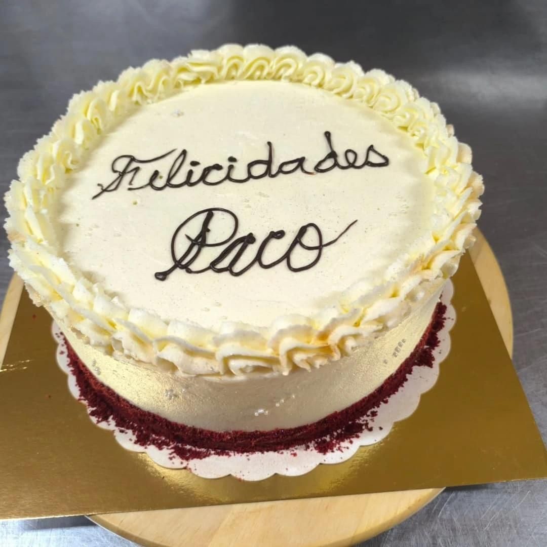 Pastel de terciopelo rojo con glaseado blanco, decorado con “Feliciades Paco” en chocolate, sobre base dorada.