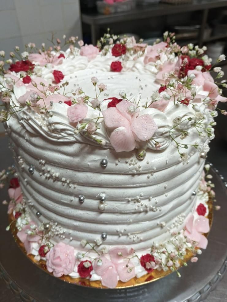 Pastel glaseado blanco, decoraciones de flores rosas y rojas, chispas plateadas, sobre una base de pastel dorada.