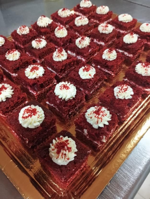 Cuadrados de pastel de terciopelo rojo cubiertos con crema y chispas dispuestos en una bandeja dorada.