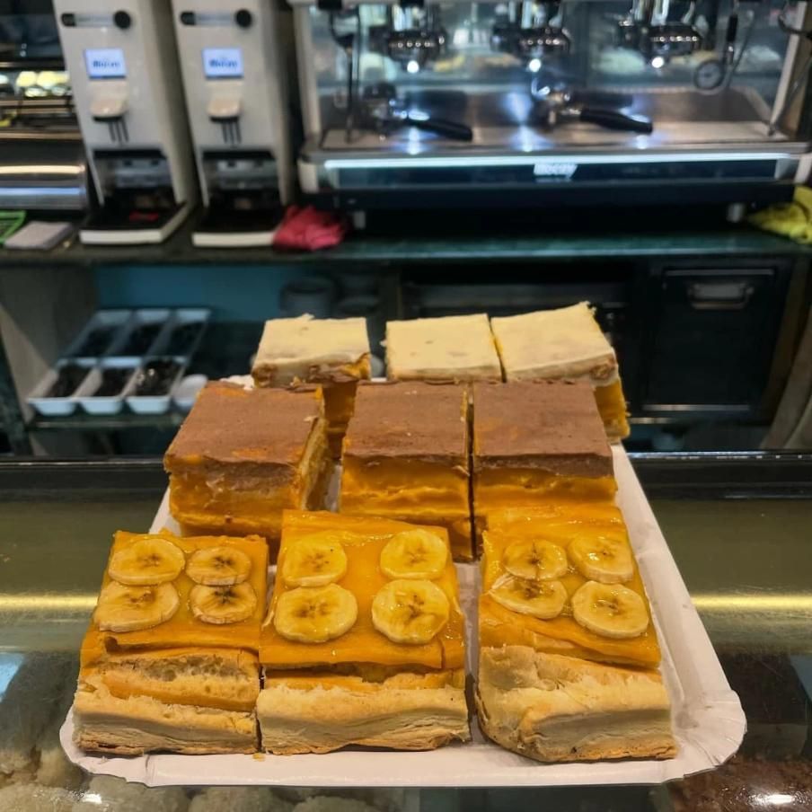 Varias rebanadas de pastel en una bandeja en un café, incluidos trozos de plátano, chocolate y cuadrados amarillos.