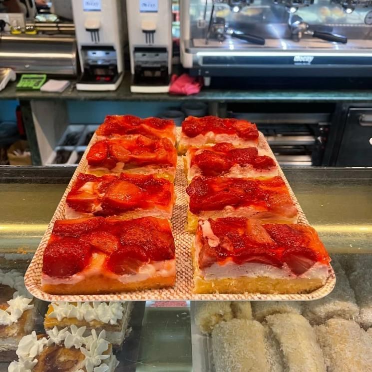 Rebanadas de tarta de fresa expuestas en una panadería, con máquinas de café al fondo.