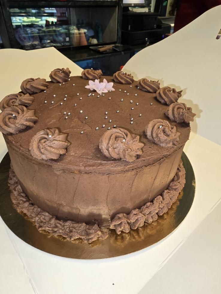 Pastel de chocolate con glaseado de chocolate, cubierto con rosetas y chispas, sobre una base de pastel dorada.