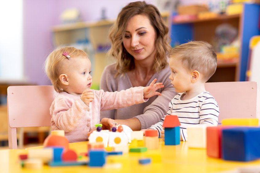 Una maestra con dos niños pequeños sentados a una mesa, jugando con bloques de colores en un aula.