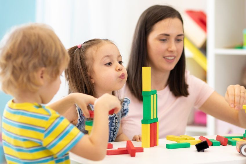 Niños y un adulto construyen bloques de colores en una mesa, con un niño apilando piezas rojas, amarillas y verdes.