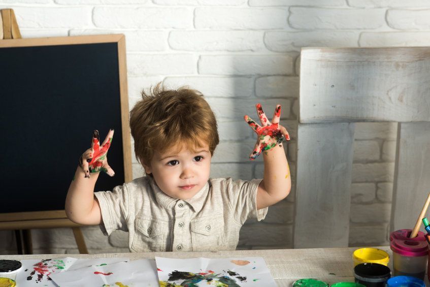 Un niño pequeño sentado en una mesa de arte, mostrando sus manos cubiertas de pintura, con pintura de colores en los dedos y las mejillas.