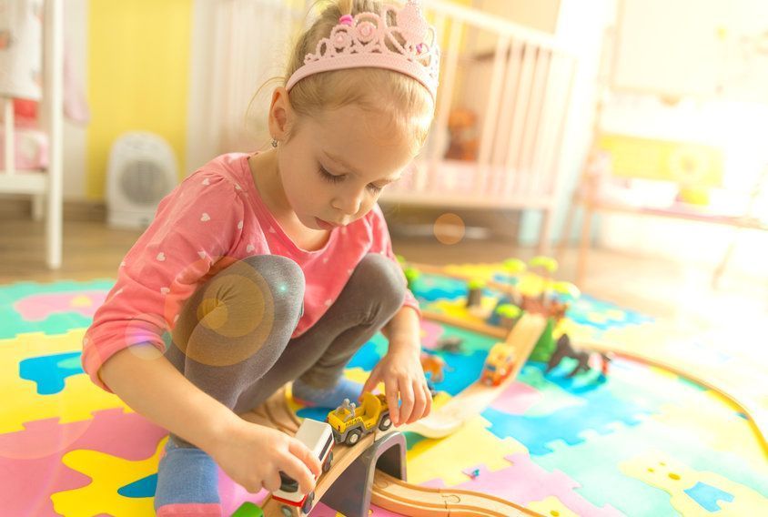 Niña con camiseta rosa y corona jugando con coches de juguete sobre una colorida alfombra de juegos.