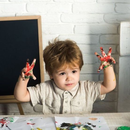 Un niño pinta en una mesa con las manos manchadas de pintura de colores, frente a una pared de ladrillos blancos y una pizarra.