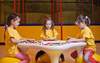 Tres niños con camisetas amarillas juegan a un colorido juego de mesa en una mesa redonda dentro de casa.