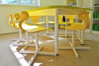 Sillas amarillas alrededor de una mesa blanca de aula en una habitación iluminada por el sol.