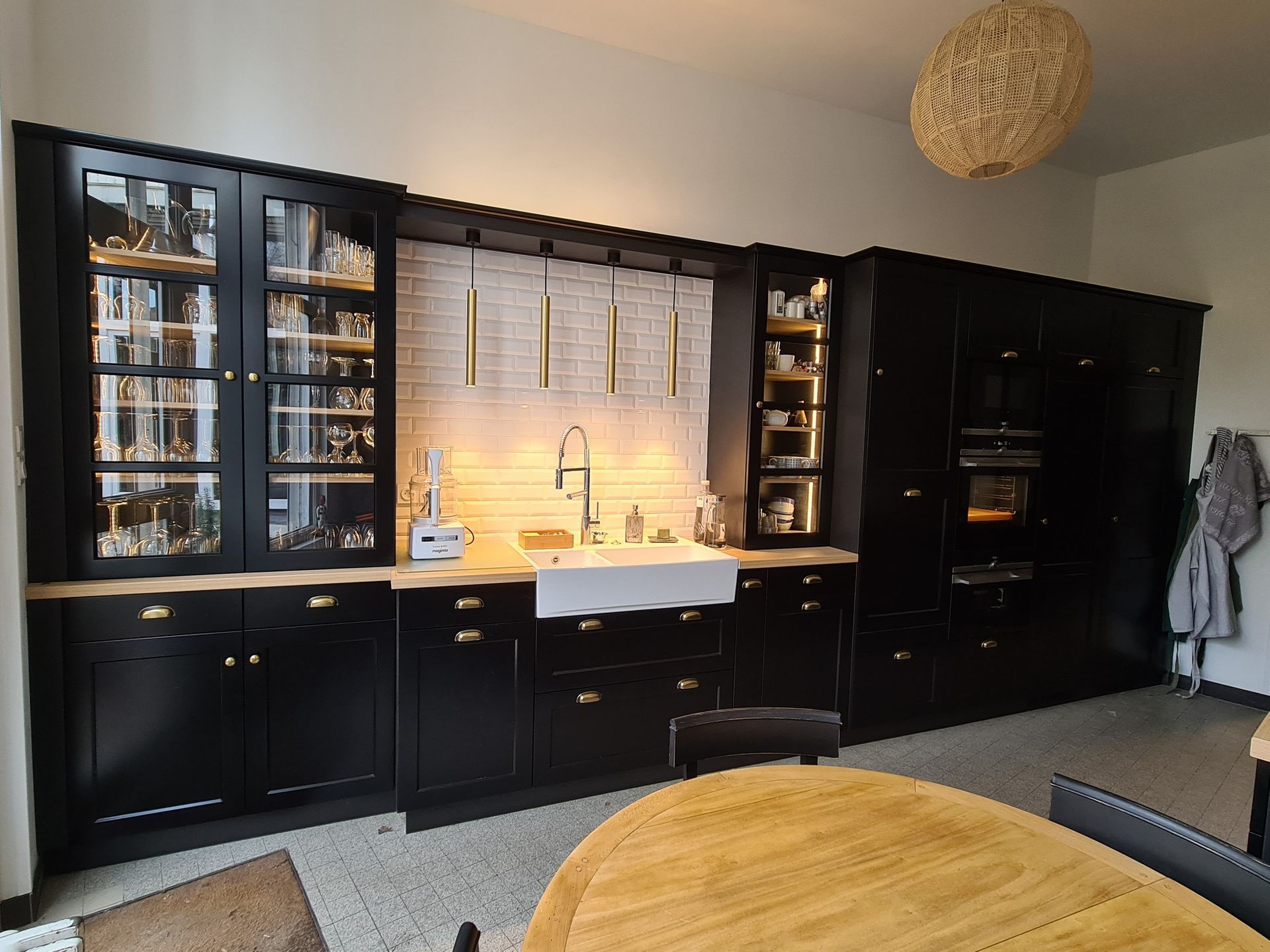 Mur de cuisine moderne noir avec placards vitrés, évier blanc sous une fenêtre éclairée et suspension lumineuse en tissu tressé