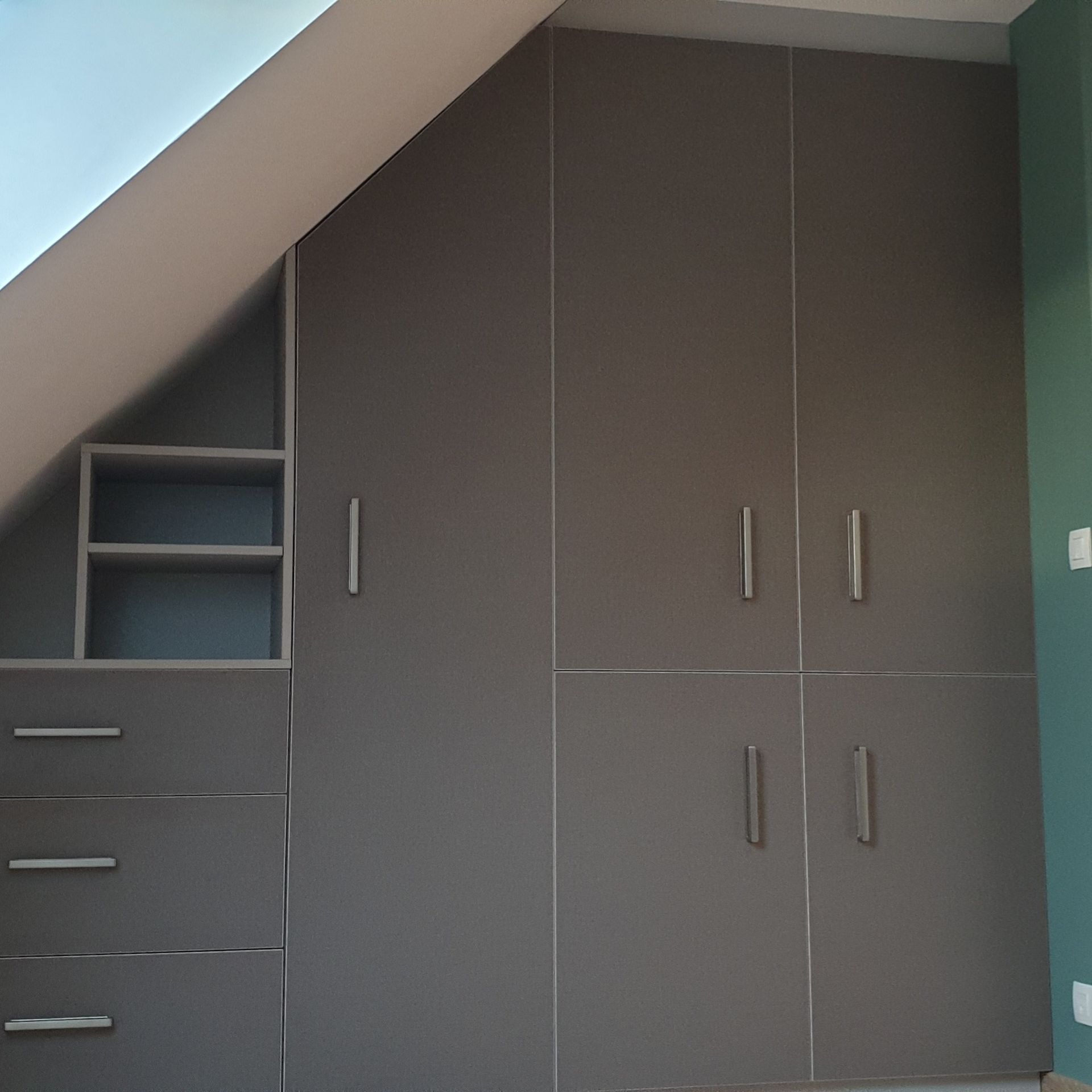 Armoire encastrée grise sous un plafond mansardé, avec tiroirs, étagères et portes de placard