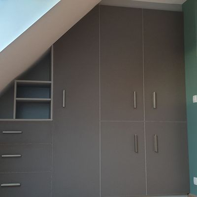Armoire encastrée grise sous un plafond mansardé, avec tiroirs, étagères et portes de placard