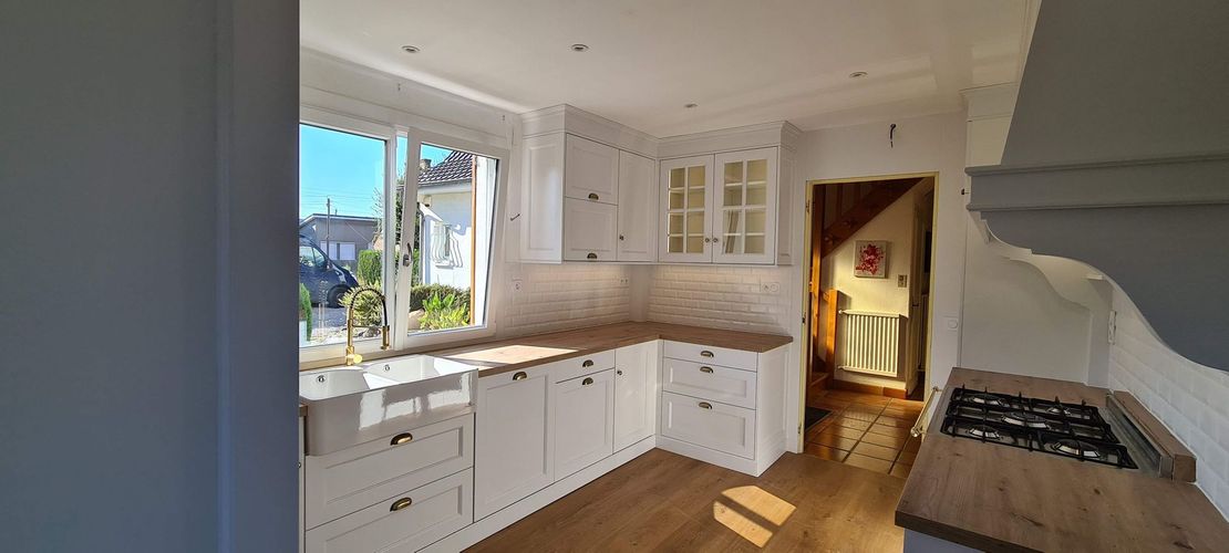 Cuisine blanche lumineuse avec parquet, fenêtre ouverte et lumière du soleil sur les plans de travail et la cuisinière.