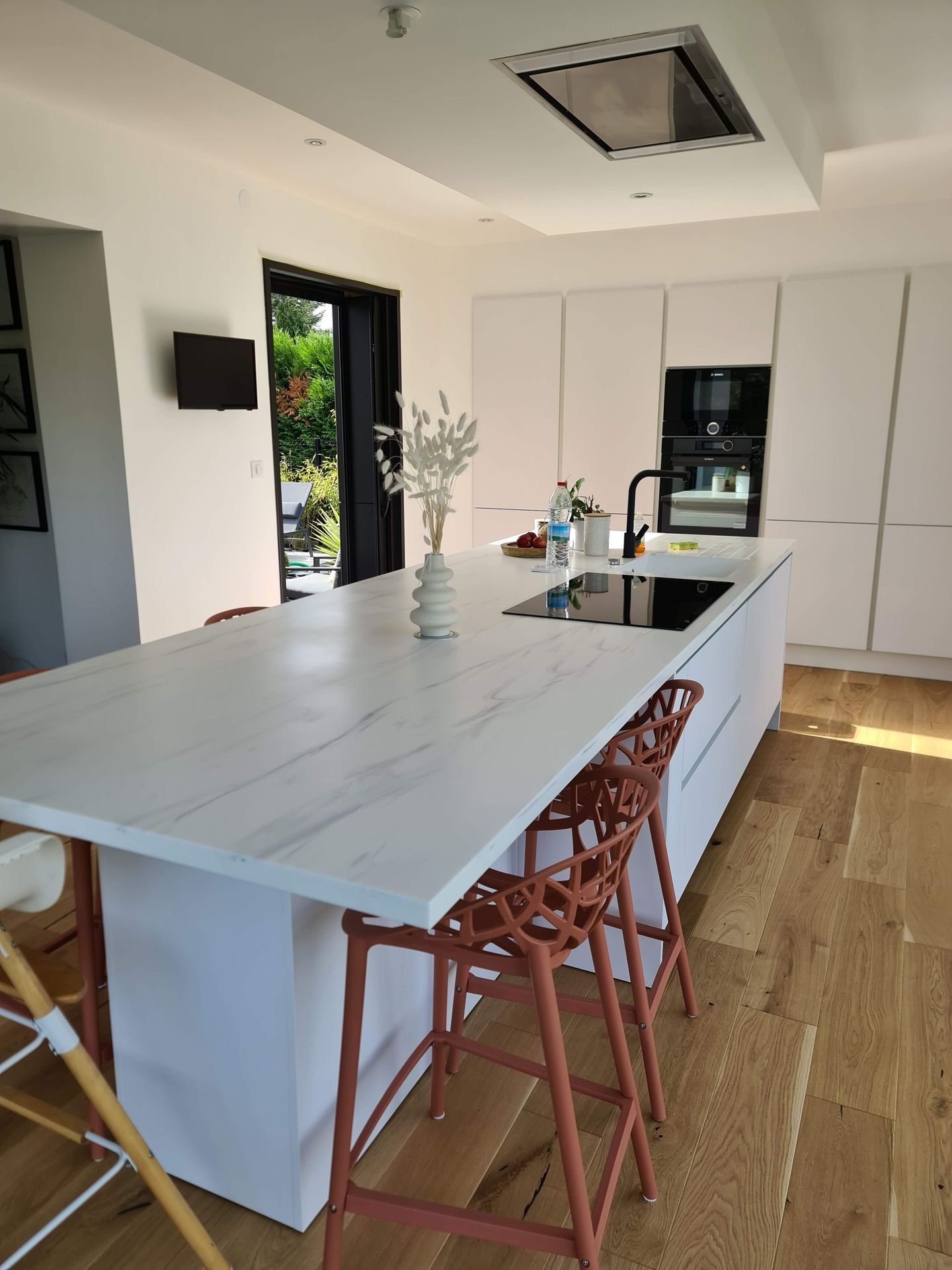 Cuisine moderne avec grand îlot blanc, tabourets en bois, touches de noir et portes vitrées ouvertes sur l'extérieur