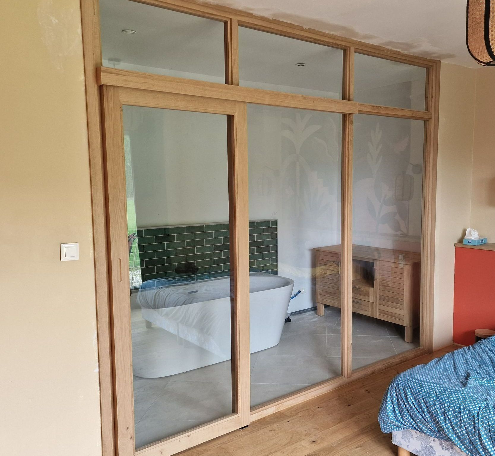 Des portes vitrées à cadre en bois donnent sur une chambre avec un lit, une baignoire, un fauteuil en osier et une armoire rouge.