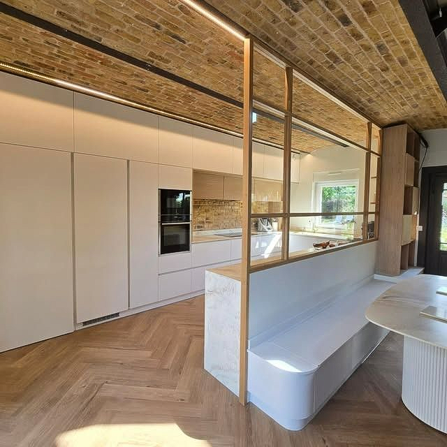 Cuisine moderne avec placards blancs, parquet, plafond en briques apparentes et cloison vitrée à côté d'un coin repas.