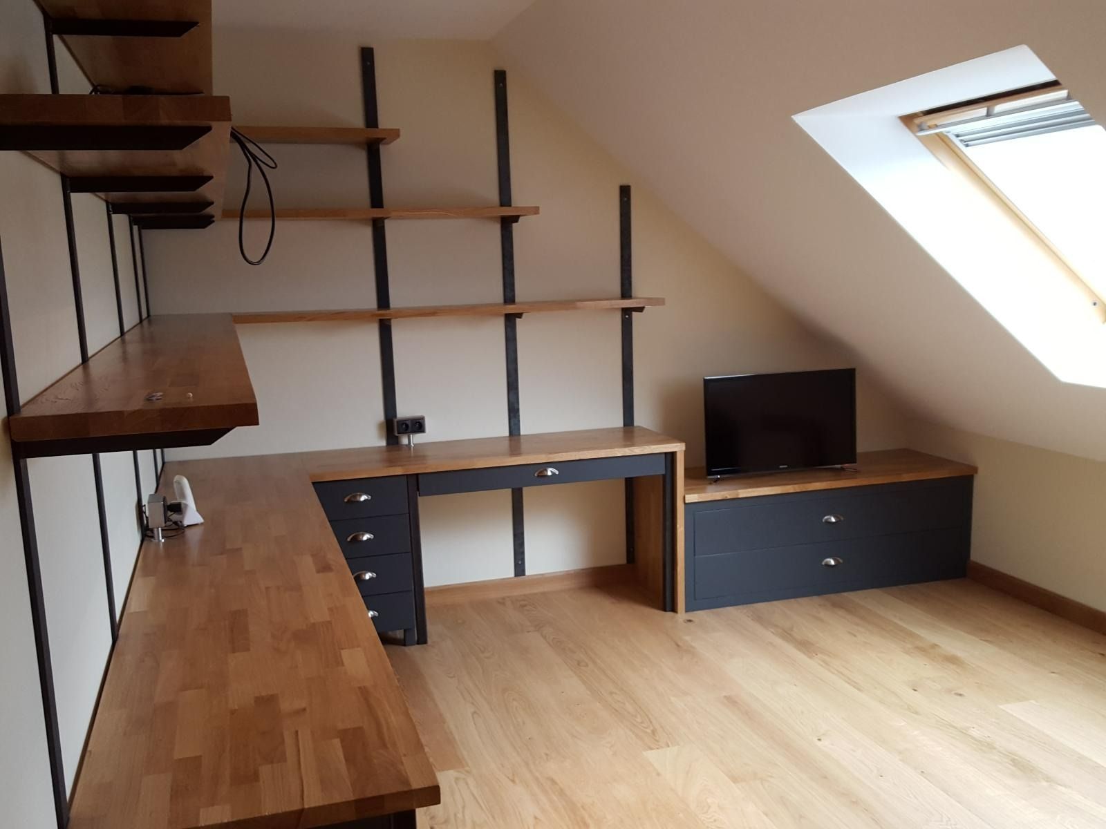 Petit bureau mansardé avec bureaux en bois, étagères, tiroirs noirs et puits de lumière sur la droite.