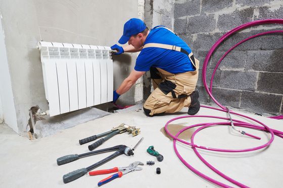 Un ouvrier installe un radiateur blanc dans une pièce en travaux, avec des outils et des tuyaux roses éparpillés sur le sol.