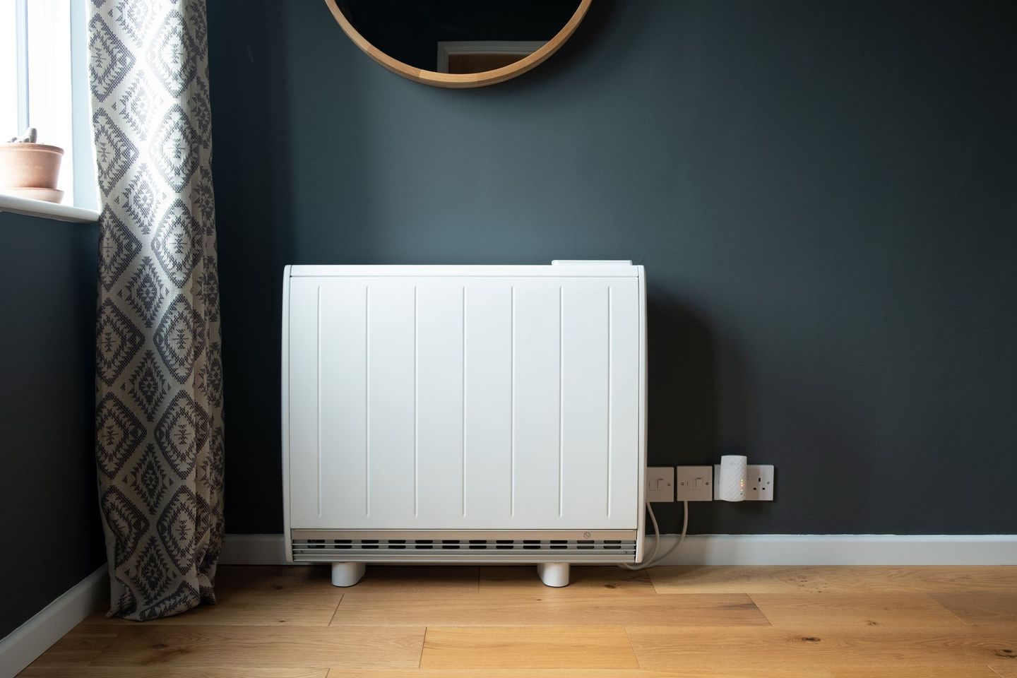 Radiateur blanc sous un miroir rond contre un mur sombre dans une pièce minimaliste avec parquet et rideaux.