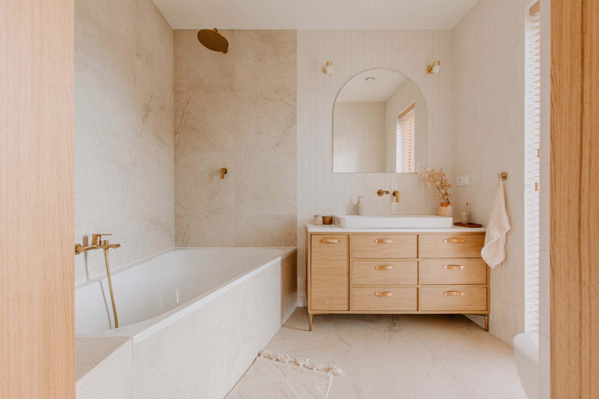 Salle de bains minimaliste beige avec baignoire, meuble-lavabo en bois, miroir arqué et serviette près de la fenêtre.
