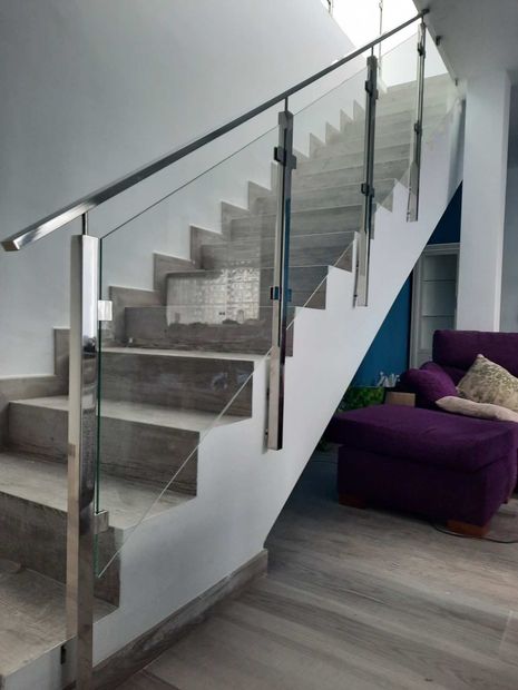 Escalera moderna con escalones de madera, barandilla de cristal y soportes metálicos sobre una pared blanca.