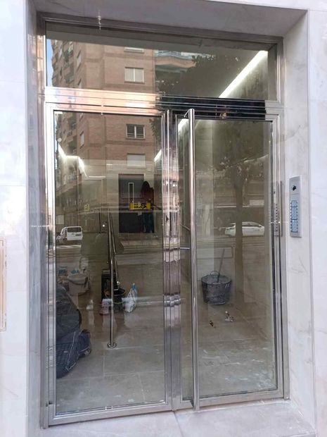 Primer plano de una puerta de acero inoxidable con tirador vertical y placa de empuje, junto a una pared revestida de vidrio.