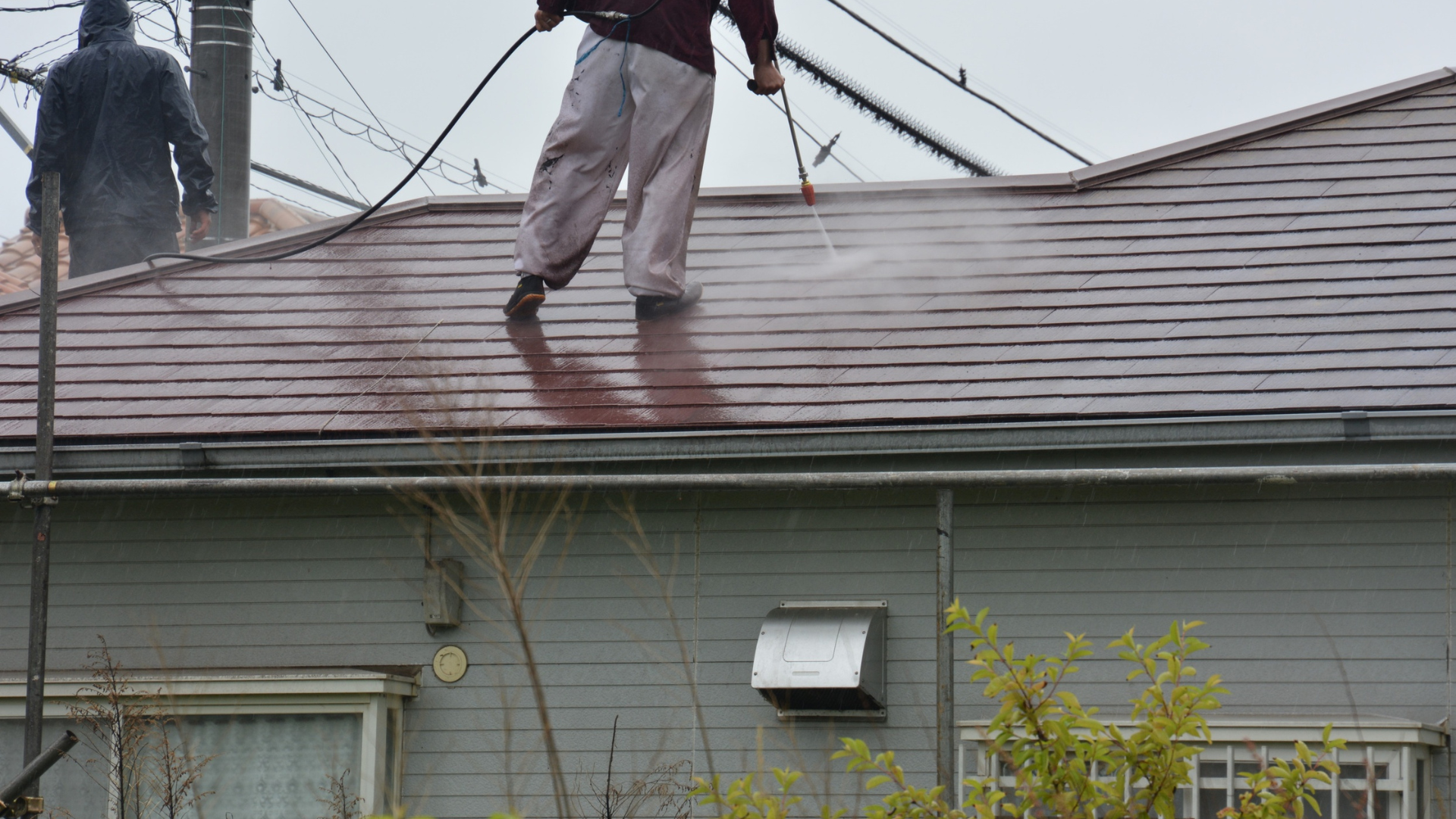 Deux personnes nettoient au nettoyeur haute pression le toit en tuiles rouges d'une maison par temps couvert.