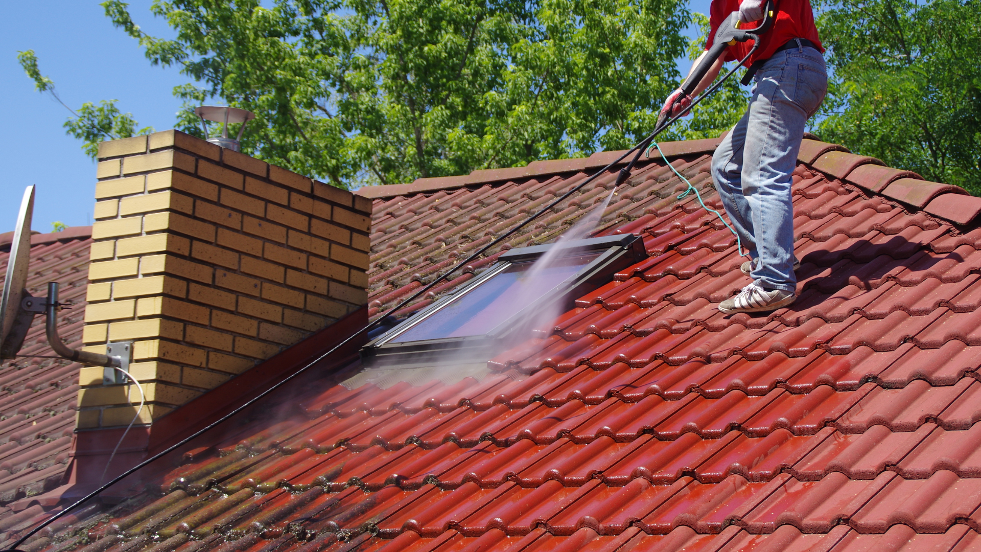 Une personne nettoie au nettoyeur haute pression un toit de tuiles rouges près d'une cheminée en briques .