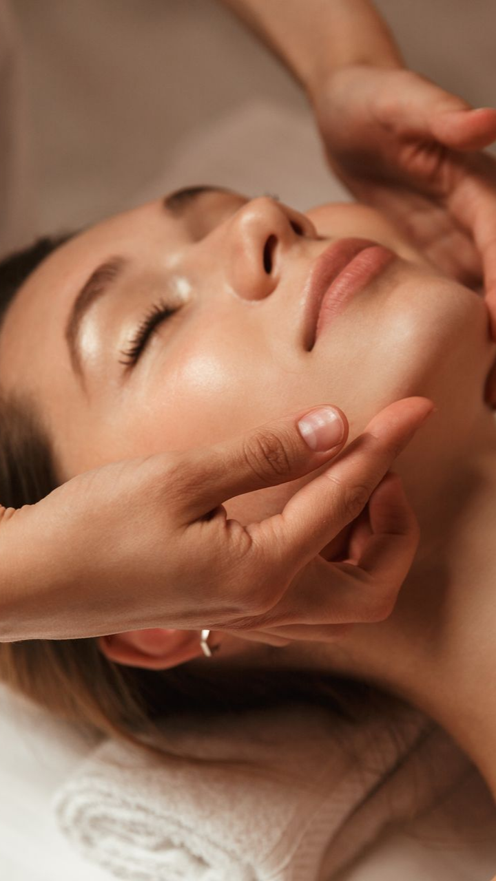 Une femme reçoit un massage du visage. Des mains effleurent son visage ; elle a les yeux fermés et est allongée.