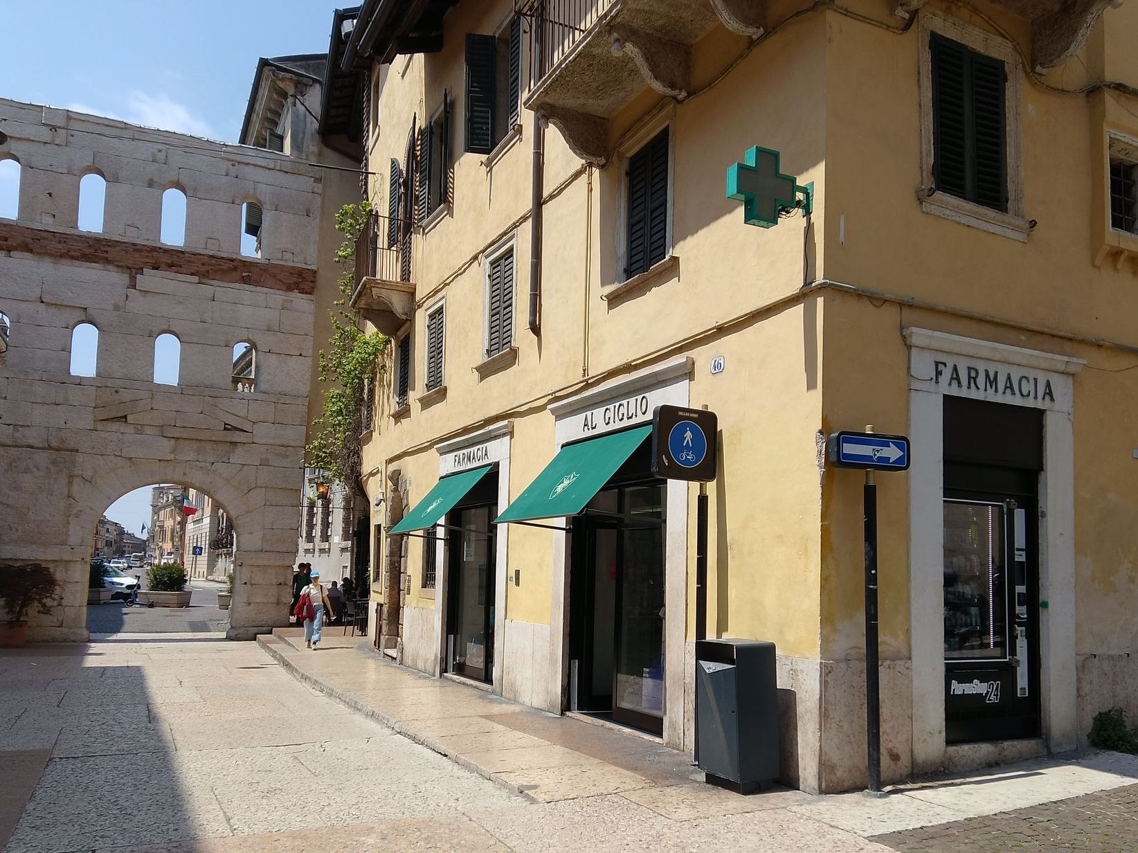 Arco in pietra accanto a un edificio giallo con l'insegna della farmacia Al Giglio e una croce verde.