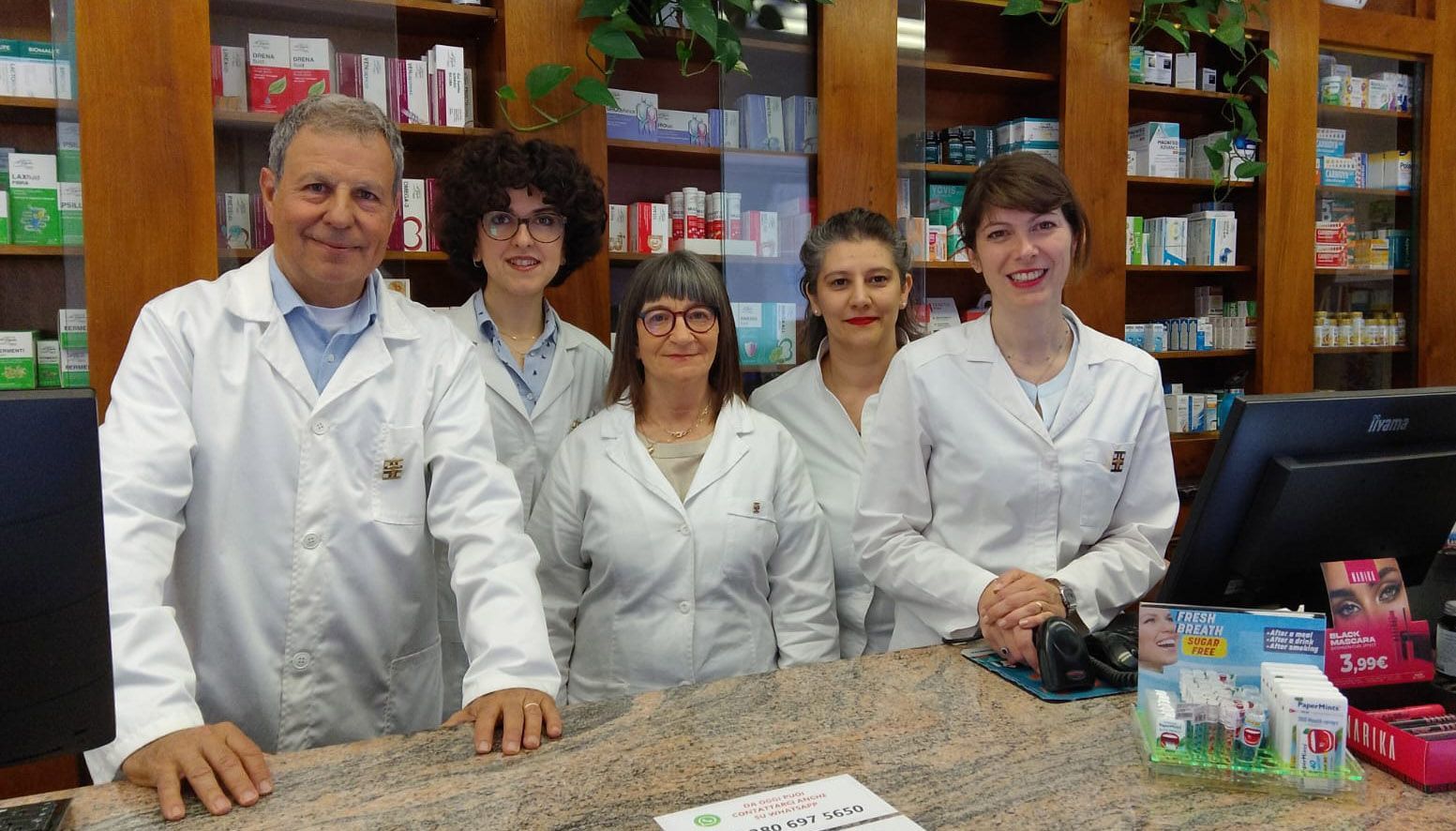 Farmacisti e personale dietro il bancone della farmacia, sorridenti, con indosso camici bianchi.