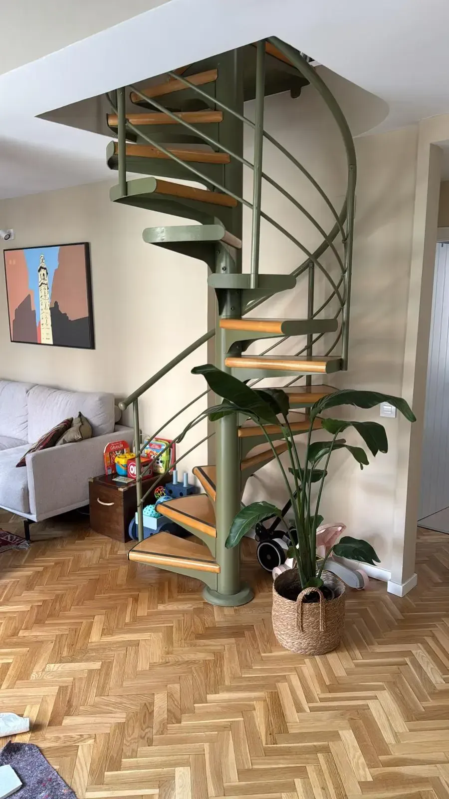 Escalera de caracol con peldaños de madera en un luminoso salón, junto a una planta y un suelo de madera en espiga.