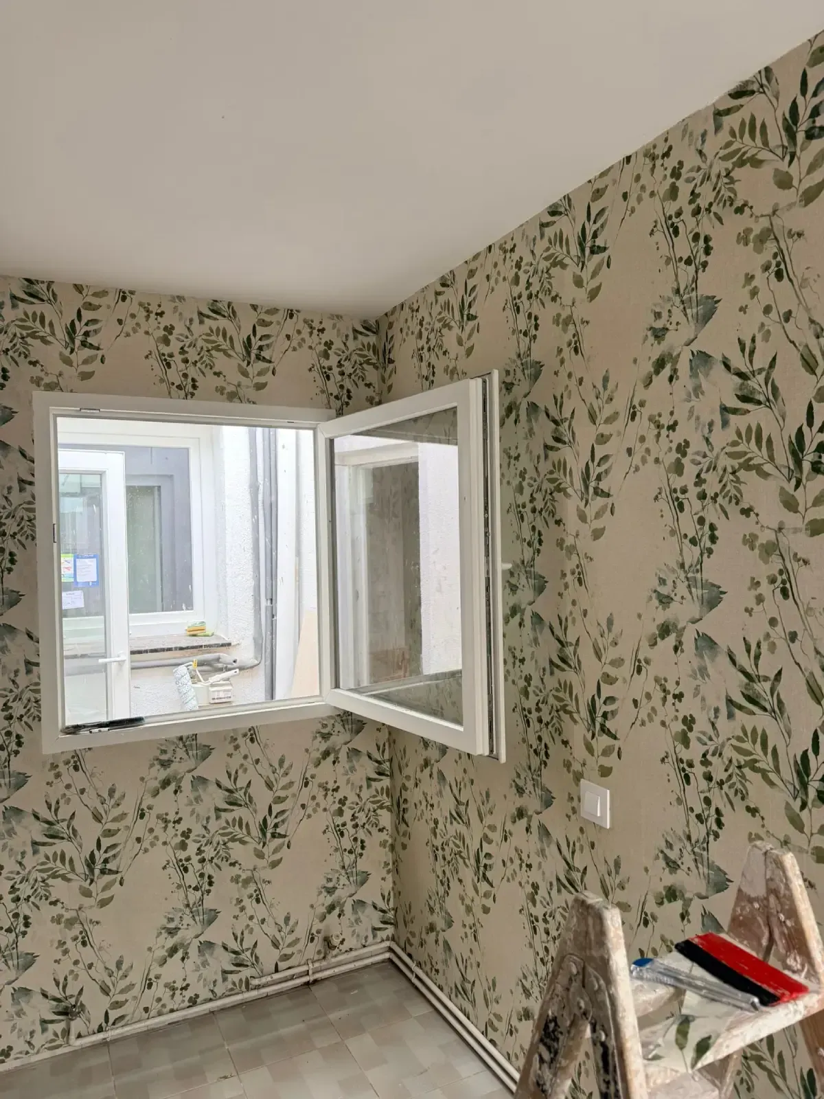 Habitación pequeña y vacía con papel tapiz floral, una ventana blanca abierta y una silla con herramientas en la esquina.