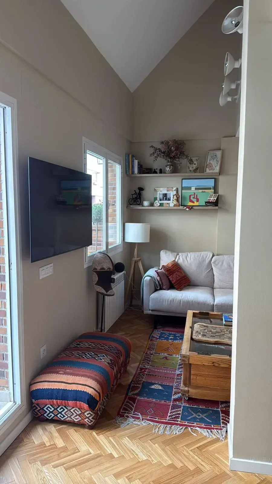Acogedora sala de estar con televisor montado en la pared, cojines junto a la ventana, sofá, alfombra