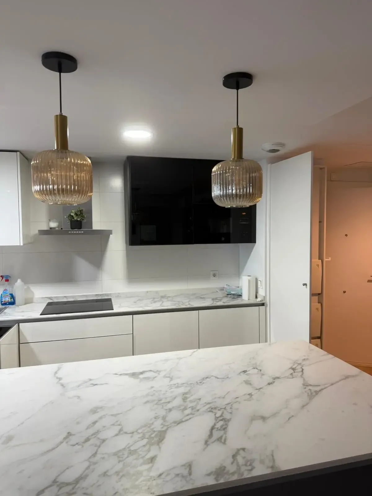 Cocina moderna con isla de mármol, armarios blancos, televisor negro y dos lámparas colgantes doradas.