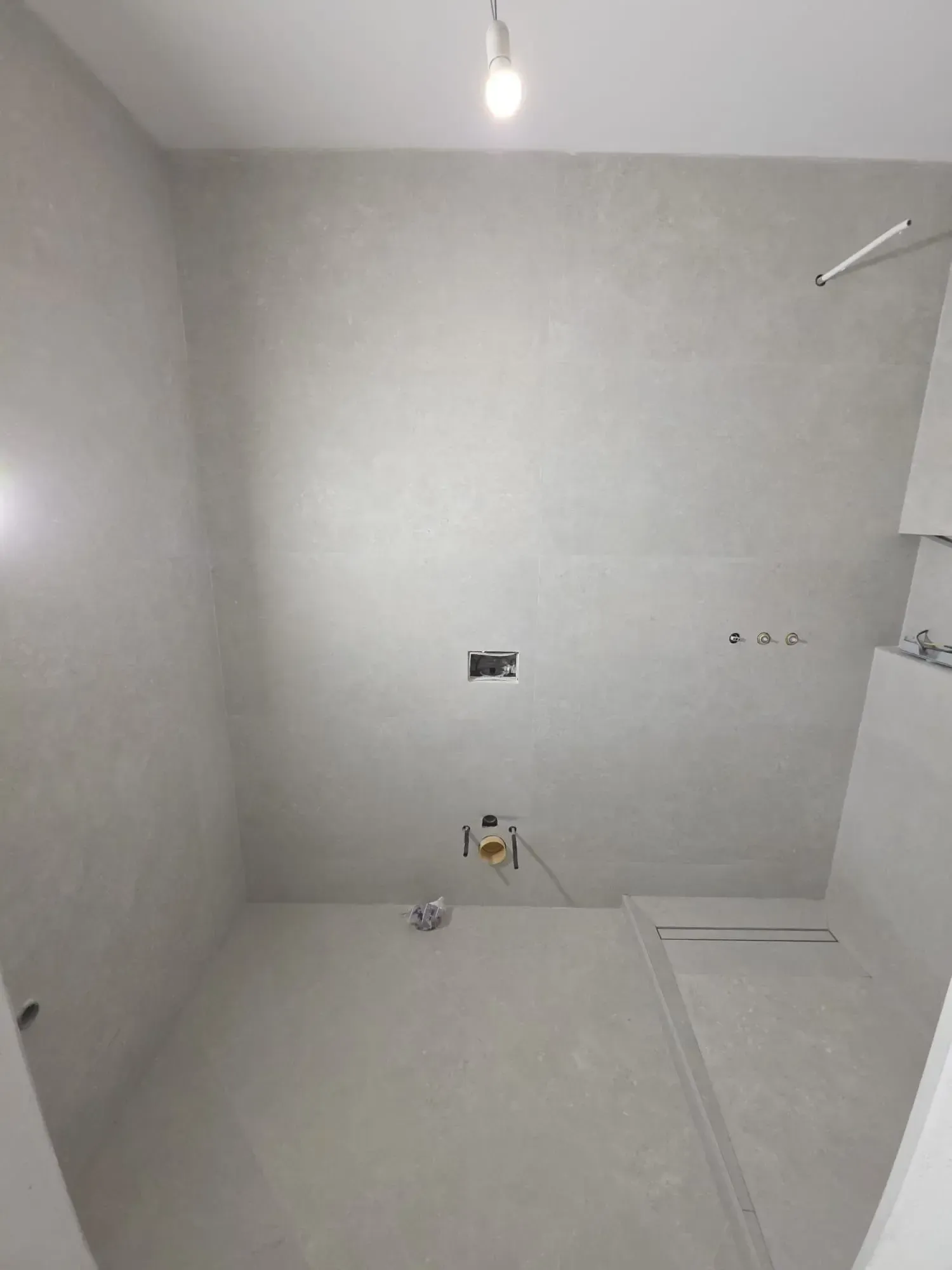 Baño sin terminar con paredes grises al descubierto, cableado expuesto y tomas de agua en el suelo y la pared.