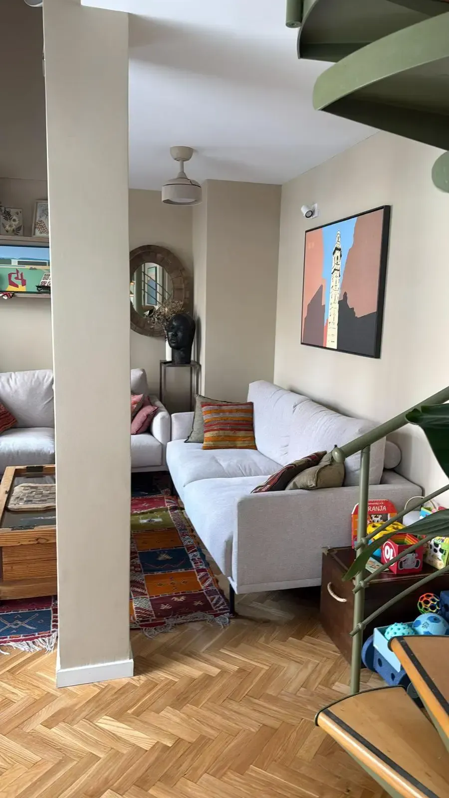 Sala de estar luminosa con sofá blanco, alfombra colorida, espejo, obras de arte y escalera de caracol.