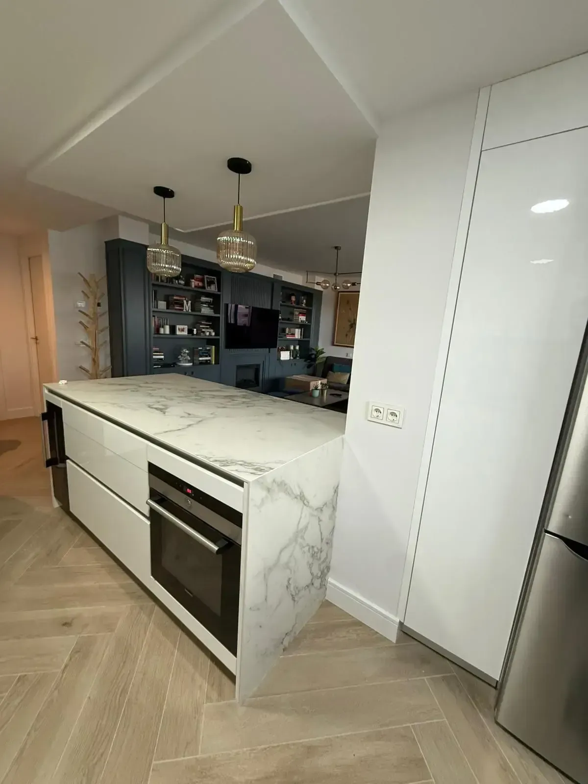Cocina moderna con isla de mármol, horno empotrado, lámparas colgantes y armarios oscuros.