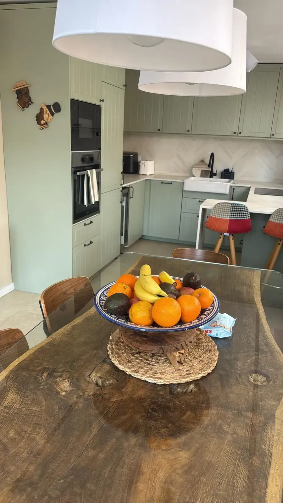Cocina con un frutero sobre una mesa de comedor, sillas rojas y armarios de color verde claro al fondo.