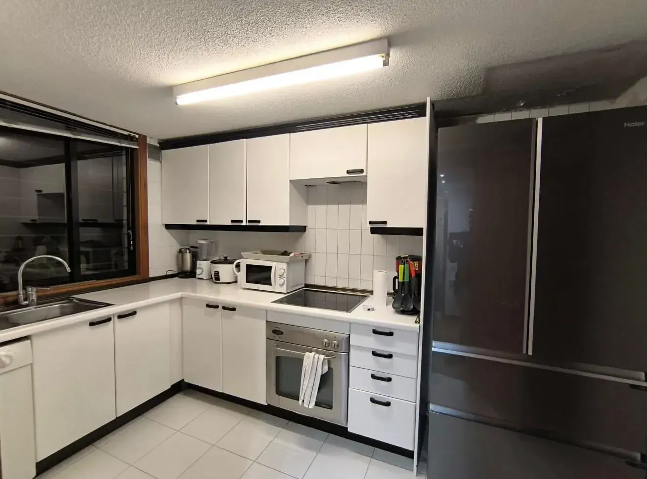 Cocina moderna blanca con electrodomésticos negros, nevera, estufa, fregadero y luz de techo fluorescente