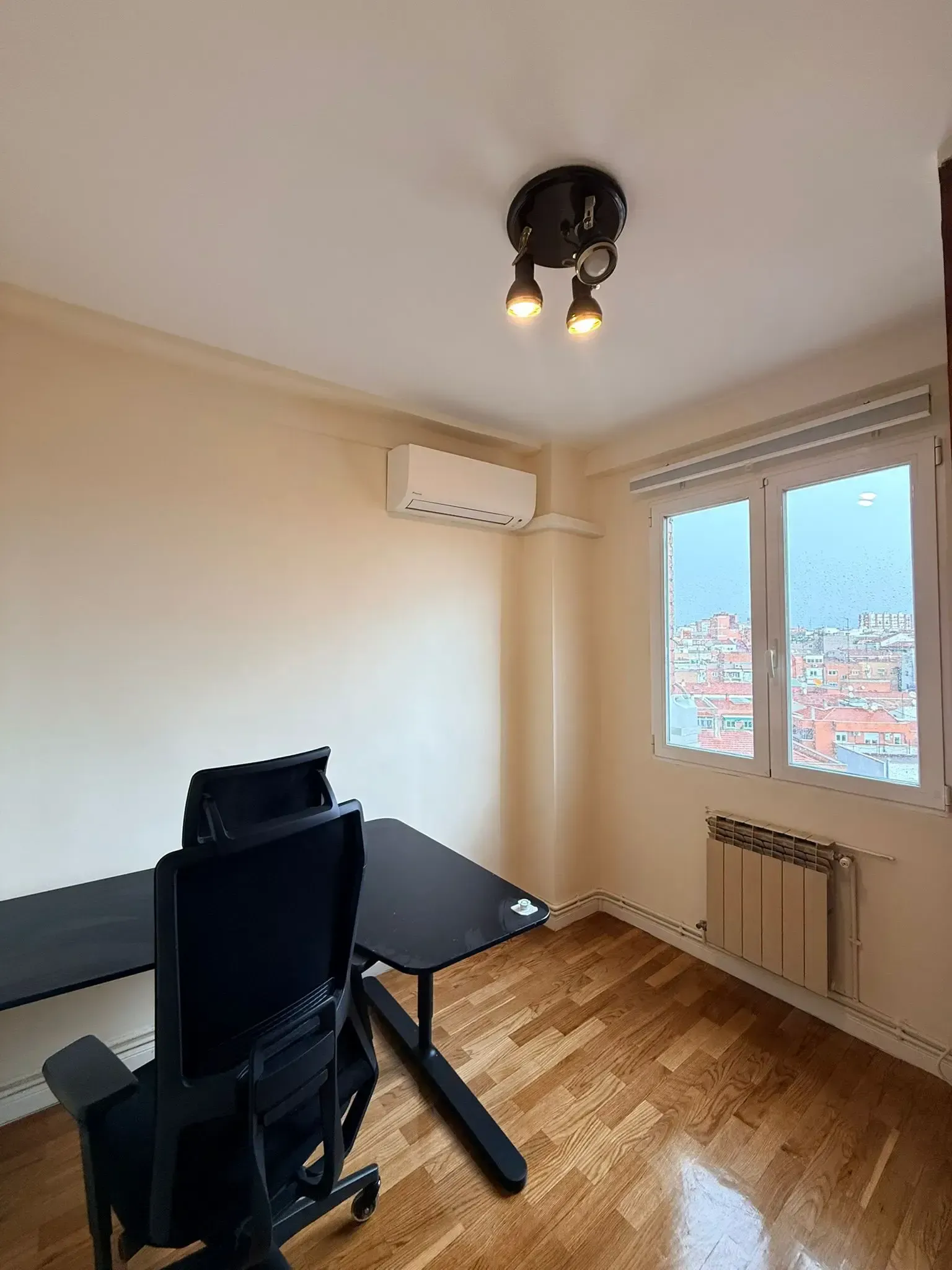 Pequeña oficina en casa con escritorio, silla, ventana, aire acondicionado mural y lámpara de techo sobre suelo de madera.