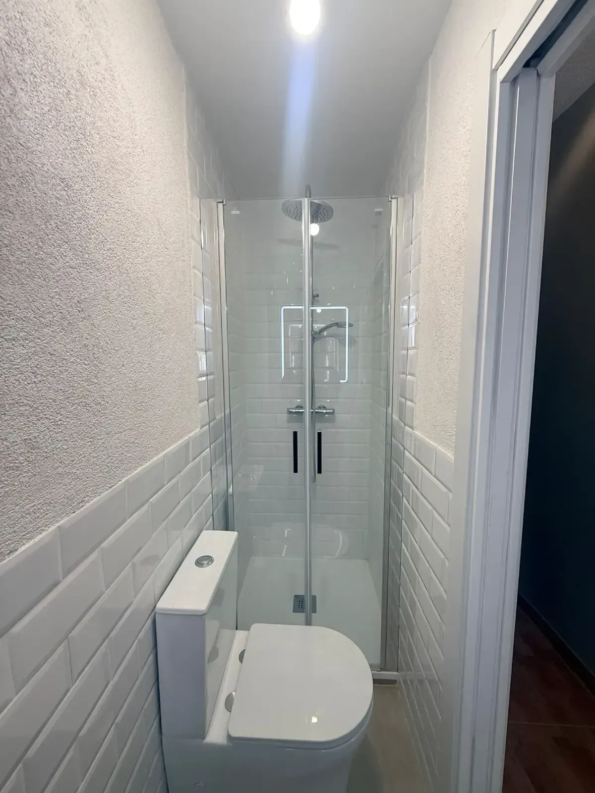 Baño estrecho con inodoro y ducha acristalada al final de un pasillo alicatado.