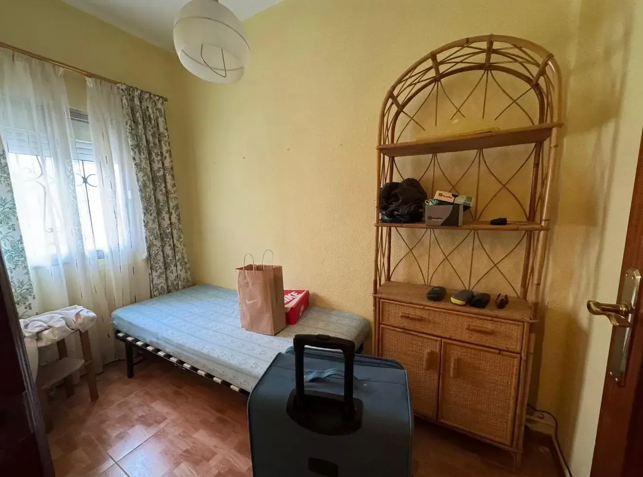 Habitación pequeña con una cama, una estantería de mimbre, cortinas en la ventana y un electrodoméstico