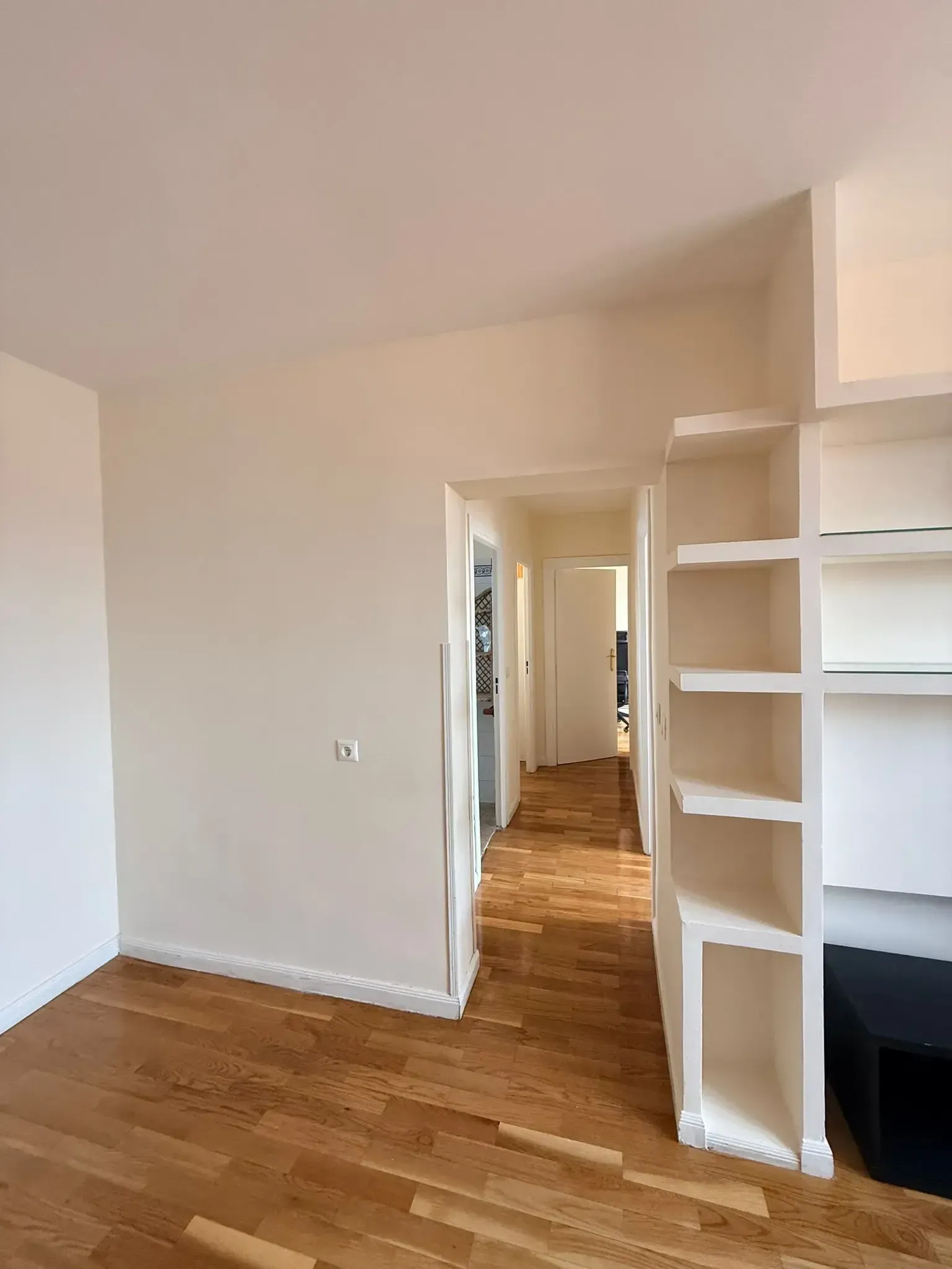 Habitación vacía con paredes blancas, suelo de madera y estanterías empotradas que dan a un pasillo.