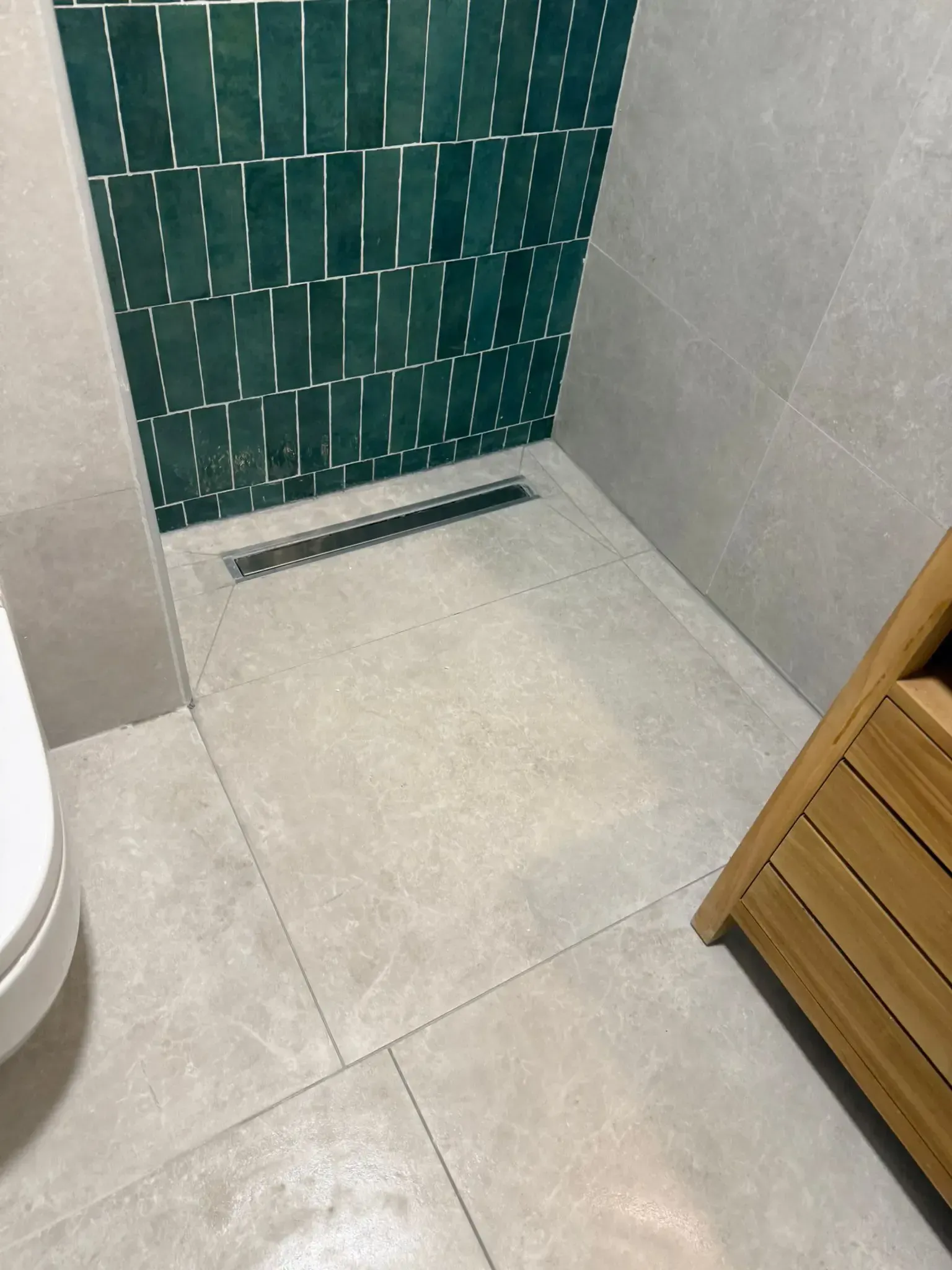 Pequeña cabina de ducha alicatada con pared de mosaico verde y desagüe lineal en el suelo.
