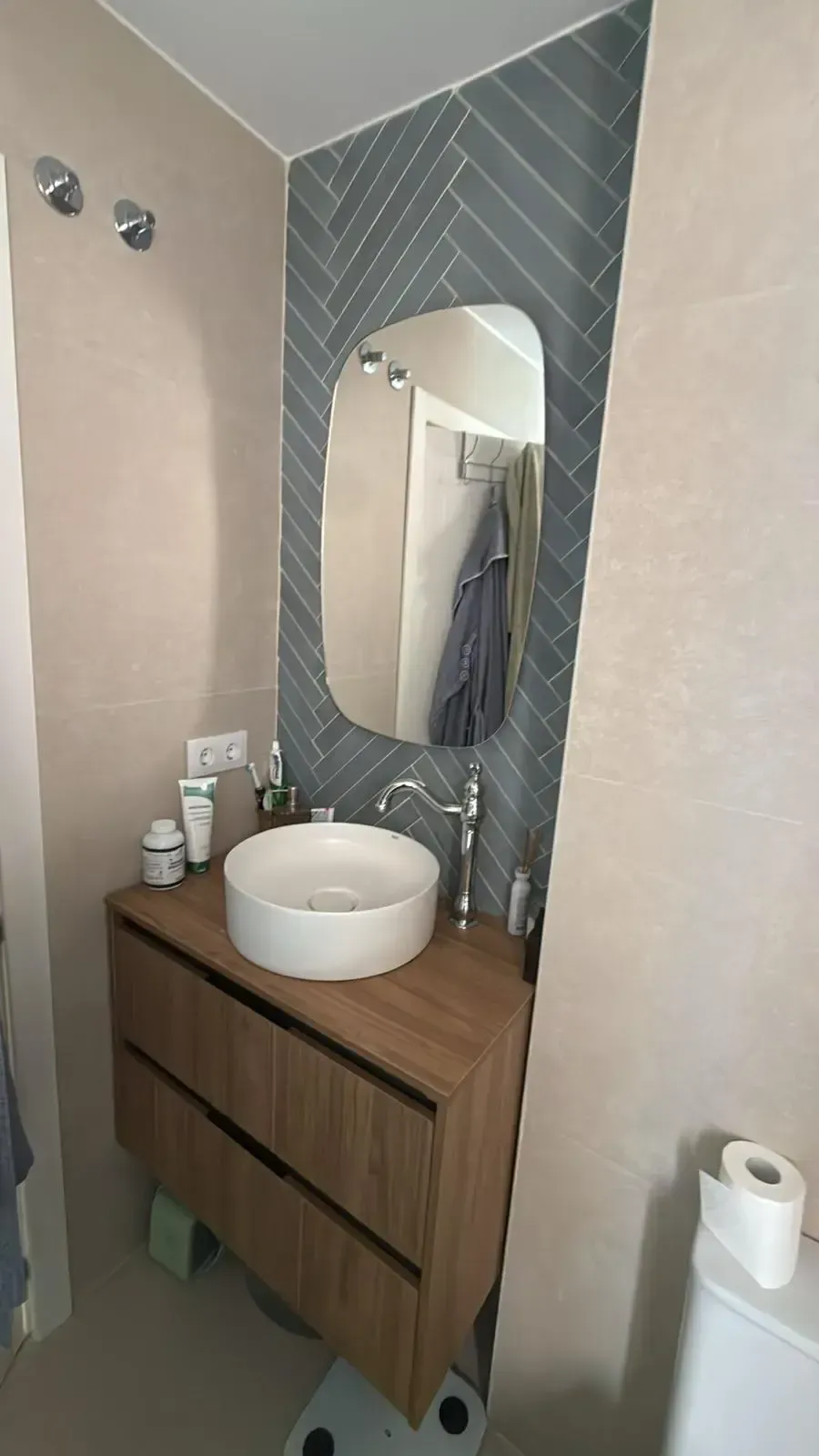 Pequeño tocador de baño con lavabo redondo, espejo, artículos de aseo y toallas colgantes sobre una pared de azulejos grises.