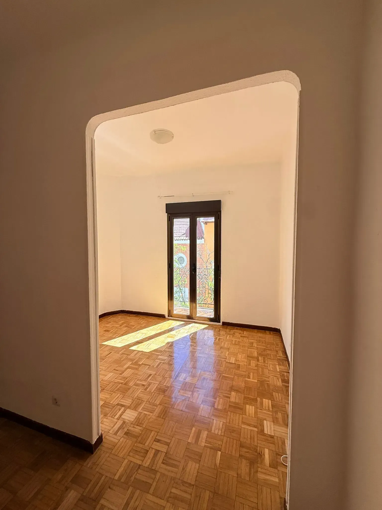 Habitación vacía con suelo de parqué, paredes blancas y una puerta de cristal que deja entrar la luz del sol.