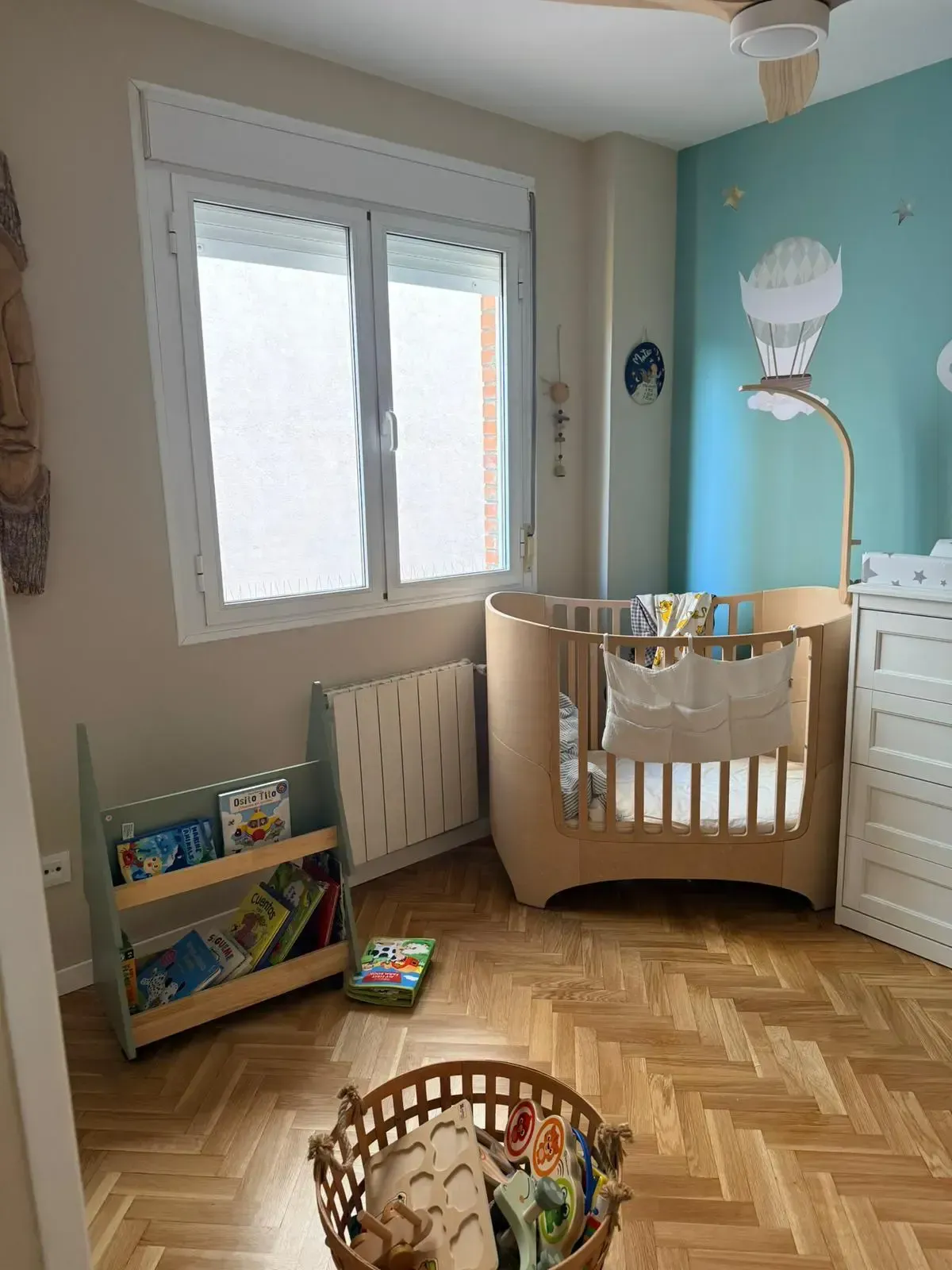 Habitación infantil con cuna, cómoda, estantería y cesta de juguetes de mimbre junto a la ventana.
