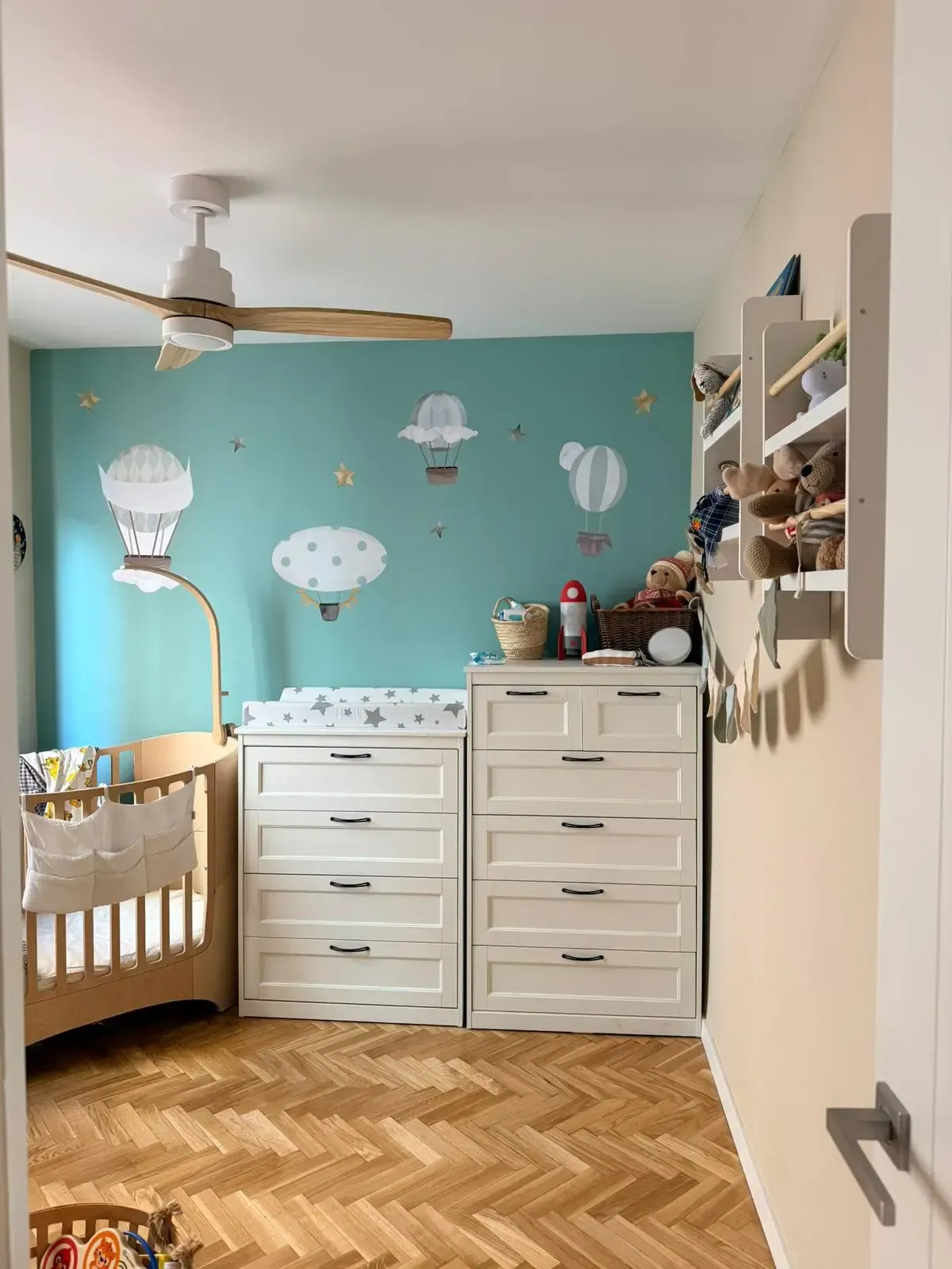 Habitación infantil con mural de globos aerostáticos color turquesa, cuna, cambiador y suelo de madera en espiga.